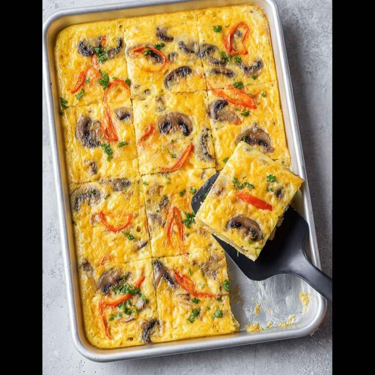 Overhead view of baked sheet pan eggs sliced into squares, topped with mushrooms, peppers, and herbs on a silver tray.
